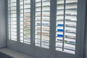 A close-up view of a window with white wooden plantation shutters partially open, allowing diffused light to enter. The scene outside is blurred but shows some buildings and a hint of greenery. For shutter installation in Tarrant County, these elegant designs add both style and functionality.