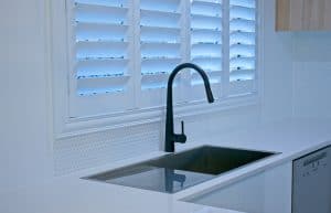 A modern kitchen with a sleek black faucet over a stainless steel sink. The countertop is white, and there is a textured white backsplash. Blue light filters through plantation shutters from Tarrant County on the window above the sink, creating a serene atmosphere. A dishwasher is visible on the right.