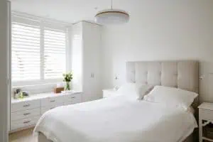 A bright, modern bedroom with white walls and furniture. A large bed with a tufted beige headboard and white bedding is centered in the room. Built-in cabinets and drawers line the wall under a window featuring plantation shutters from Tarrant County. A white pendant light hangs from the ceiling.