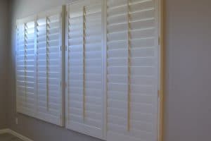 A set of four white plantation shutters covering a wide window in Tarrant County. The shutters have horizontal slats and are hinged, allowing them to open and close. The room has grey walls and a wooden floor, with natural light filtering through the gaps between the slats.