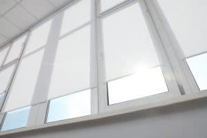 A series of large, white-framed windows with roller blinds partially pulled down. Sunlight is peeking through one of the windows, creating a bright, serene atmosphere. The window ledge and walls are also white, contributing to the minimalist aesthetic often seen in homes after blinds installation in Tarrant County.