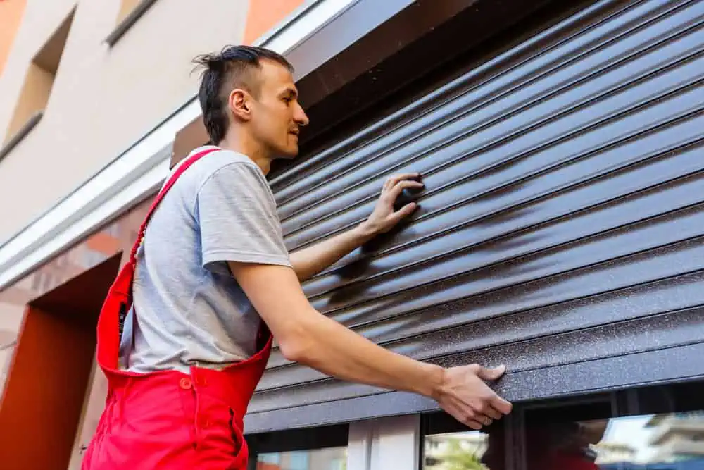 A person in a gray t-shirt and red overalls rolls up an exterior metal shutter on a building. The individual uses both hands to lift the black shutter, with part of the building and windows visible in the background.