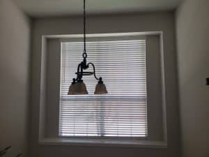 A pendant light with two lampshades hangs in front of a large window covered by white blinds. The room, dimly lit and shadowed, offers a serene ambiance, with subtle plays of light and shade that dance across walls and ceiling.