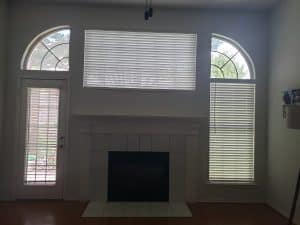 A living room features a fireplace centered on a white wall, above which is a large window with closed shades. Flanked by two smaller arched windows also fitted with shades, the space includes a door to the left with a glass panel and matching blinds.