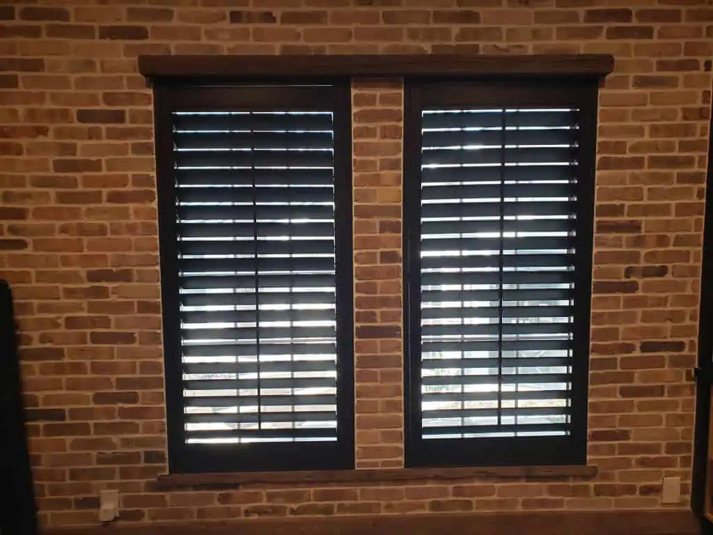 Two tall windows with closed black shutters are set in a brick wall, resembling dark shades against the mix of light and dark bricks. Sunlight filters through the slats, adding a soft glow to the textured façade.