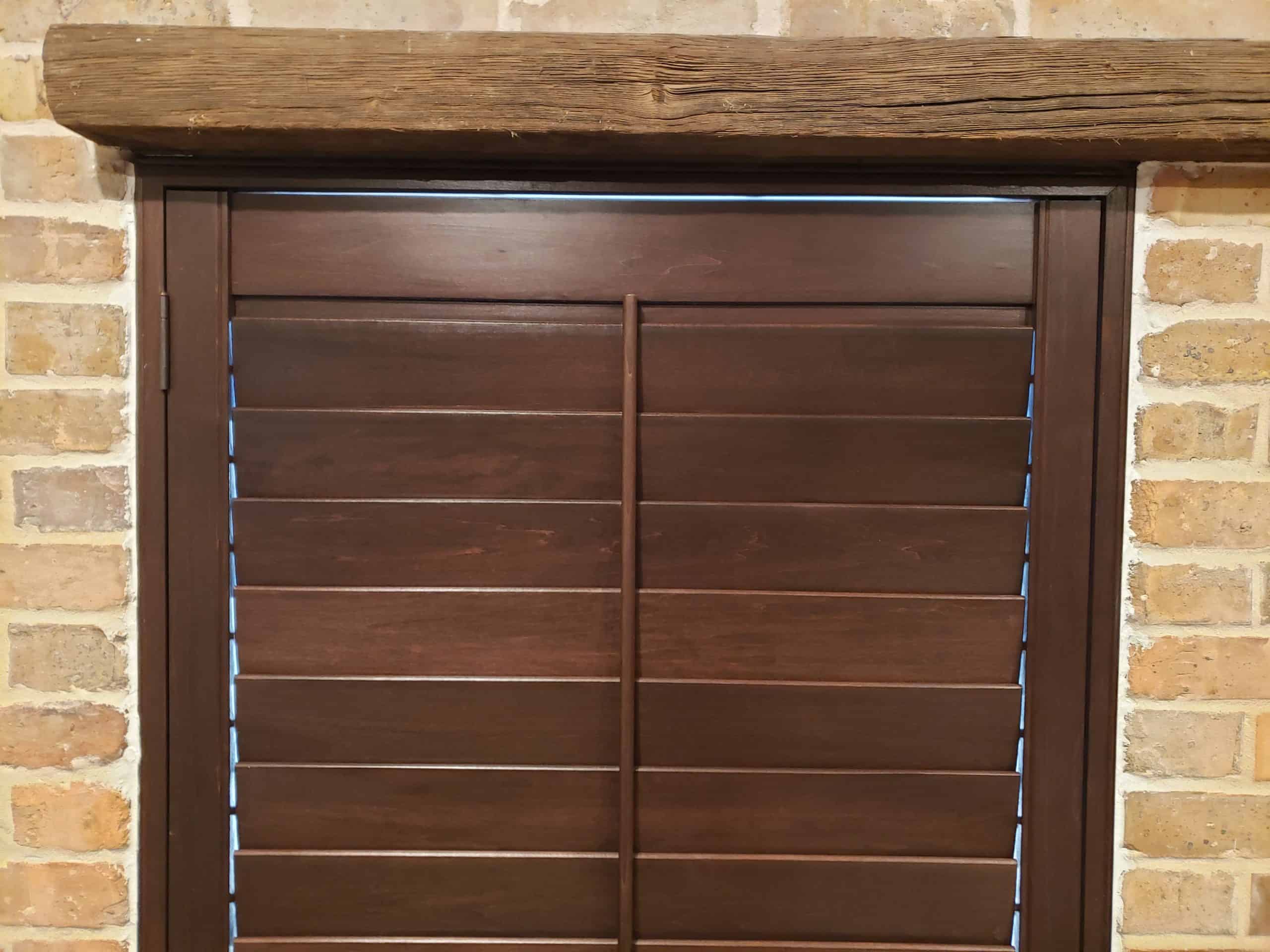 A wooden window shutter with horizontal slats is set against an exposed brick wall, much like classic shades. The shutter is framed by a thick wooden beam at the top. The natural tones of the wood and brick create a rustic appearance.