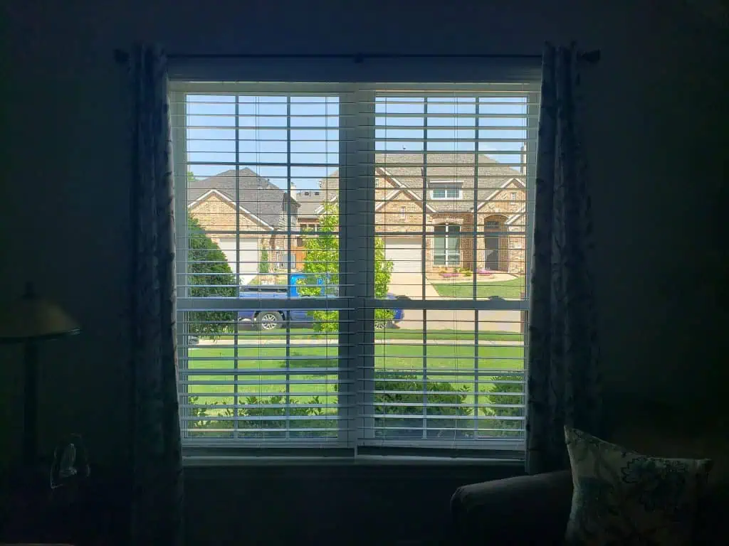 A view through a window with white shades partially open reveals a suburban neighborhood, where the street is lined with green lawns and brick houses. A blue car rests in the driveway of a house across the door, completing this peaceful scene.