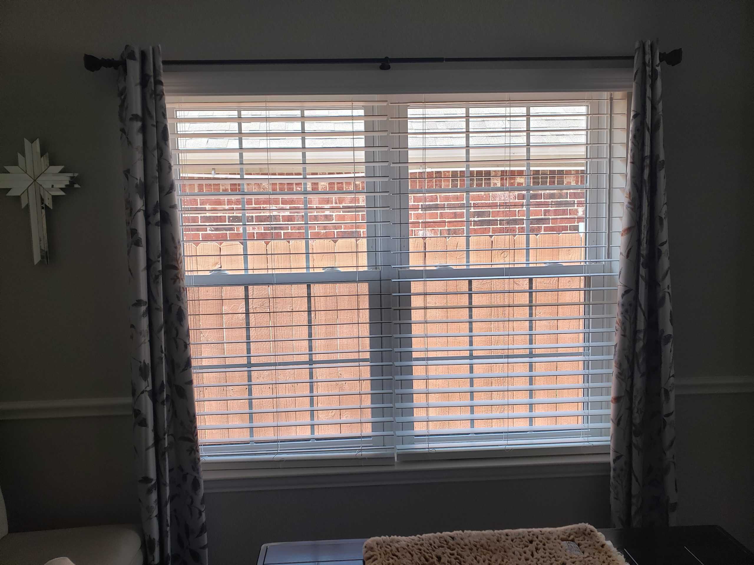 A window with white blinds partially open reveals a view of a brick wall outside. Dark patterned curtains frame the scene, while shades add a touch of elegance. A light-colored blanket is partially visible on the wooden surface below, offering a cozy contrast.
