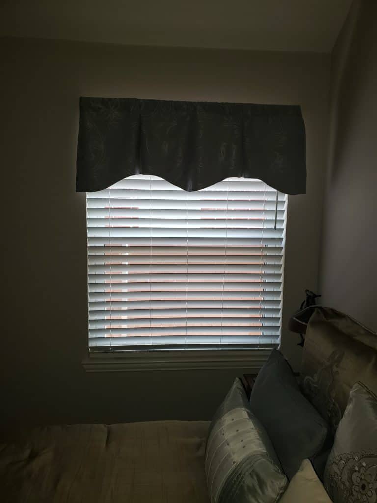 A window with closed blinds and a dark valance takes center stage, flanked by elegant doors. Below lies a bed adorned with decorative pillows in shades of gray and blue. Gentle light filters through the blinds, casting a serene atmosphere throughout the room.