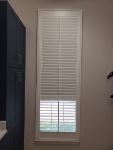 A tall, narrow window with white horizontal blinds, partially open, allows light to reveal a dark cupboard with silver handles to the left and a section of our About Us-inspired white wall-mounted storage rack to the right.