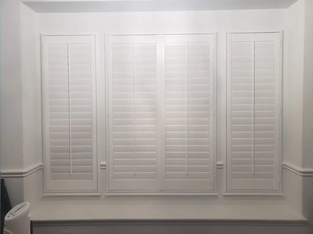 White wooden plantation shutters adorn three large windows in a pristine room, their horizontal slats firmly closed, allowing no light to seep through. Below the shutters, a white window ledge adds to the room's elegance. These classic shades bring a touch of timeless charm to any decor.