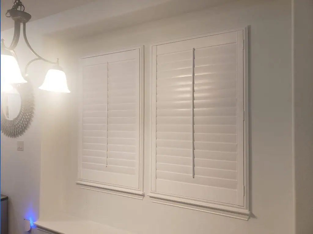 Two white plantation shutters on a wall with closed slats resemble elegant shades. A nearby chandelier casts light, illuminating the shutters and surrounding area. A decorative door-shaped wall mirror is partially visible to the left.