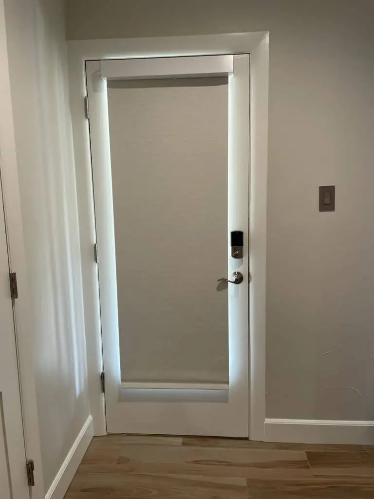 A white door with a light gray roller shade elegantly covers the window, blending seamlessly into the light gray walls. The door features a sleek silver handle and a digital lock. Light wood floors complement the space, while a convenient light switch rests nearby.