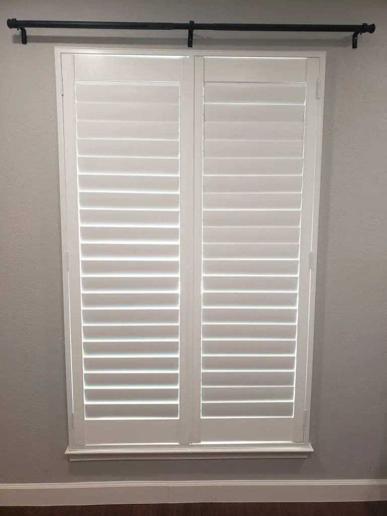 A window with closed white plantation shutters and sleek shades adorns a room with light gray walls. A black curtain rod is installed above the window, complementing the dark wood floor gracefully.