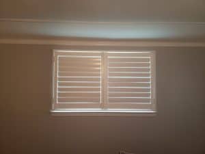 A wall with a rectangular window covered by closed white blinds. Soft light filters through the gaps, creating stripes on the blinds. The wall is painted a neutral color, and a small section of the ceiling is visible at the top.
