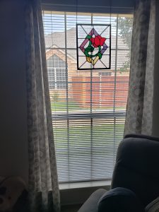 A window with vertical blinds partially open, showing a stained glass piece with a rose design hanging at the top. Patterned curtains frame the window. Outside, a red brick house and green lawn are visible. A blue cushioned chair is in the foreground.