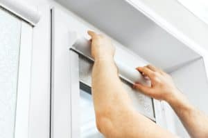 A person’s hands are installing or adjusting a white roller window blind on a window with a white frame. The image focuses on the upper part of the window and the person's arms.