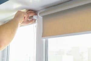 A person’s hands are installing or adjusting a beige roller blind on a window, pulling the chain to operate the blind. The scene is indoors with natural light coming through the window.