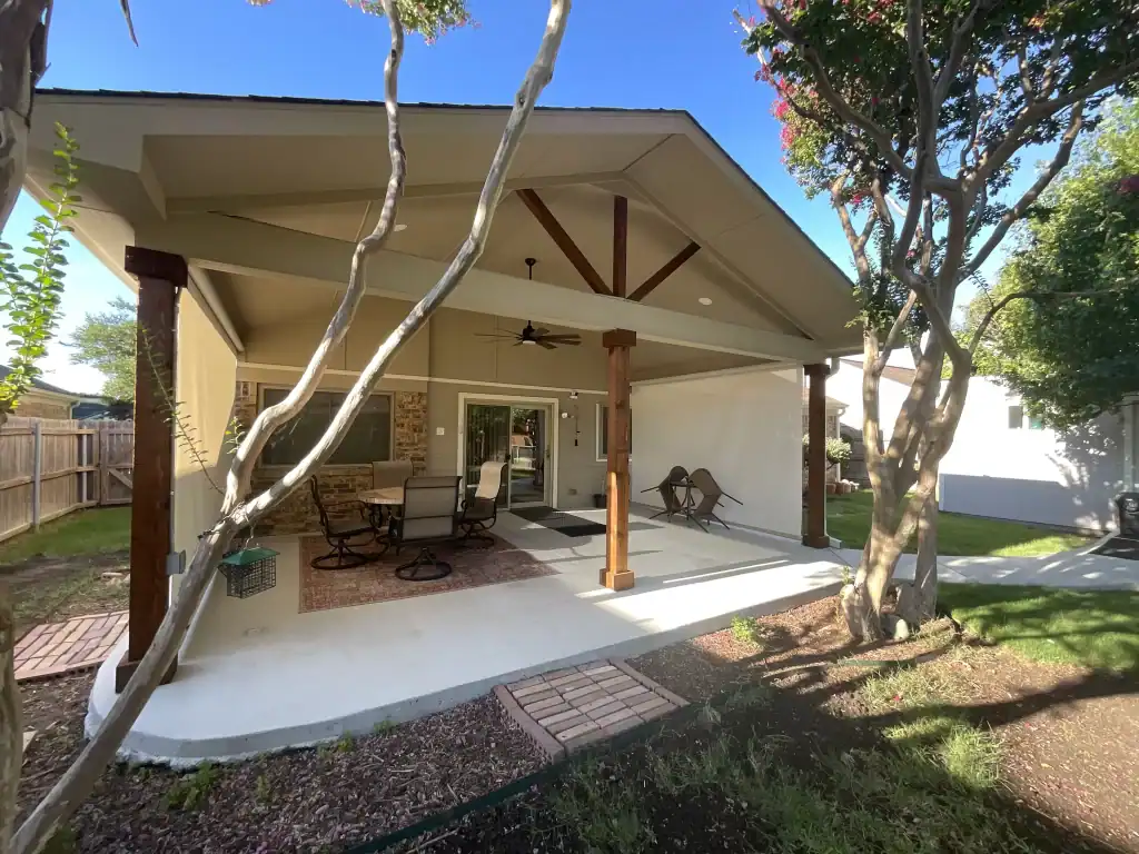 A covered backyard patio with wooden posts, outdoor furniture, ceiling fan, and two trees in the foreground. The patio opens to a garden and a fenced yard.