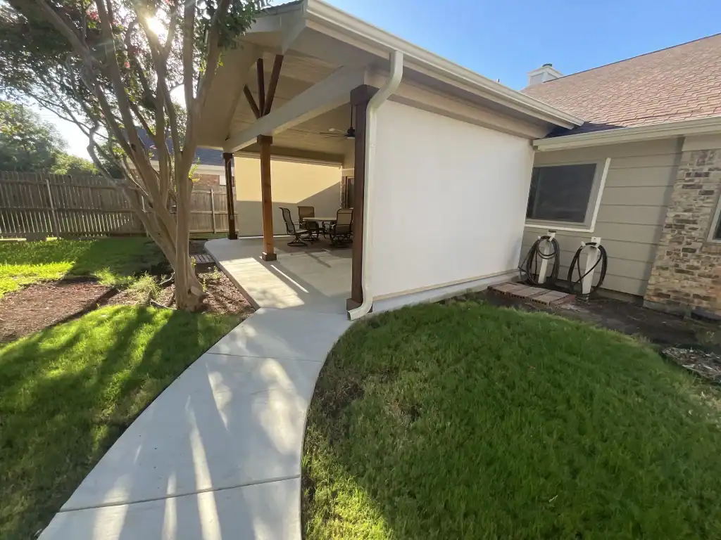 A concrete path curves through a green lawn to a covered patio with outdoor seating. The patio, attached to the house and partly enclosed by a white privacy screen, features stylish shades and doors. Sunlight casts shadows from nearby trees.