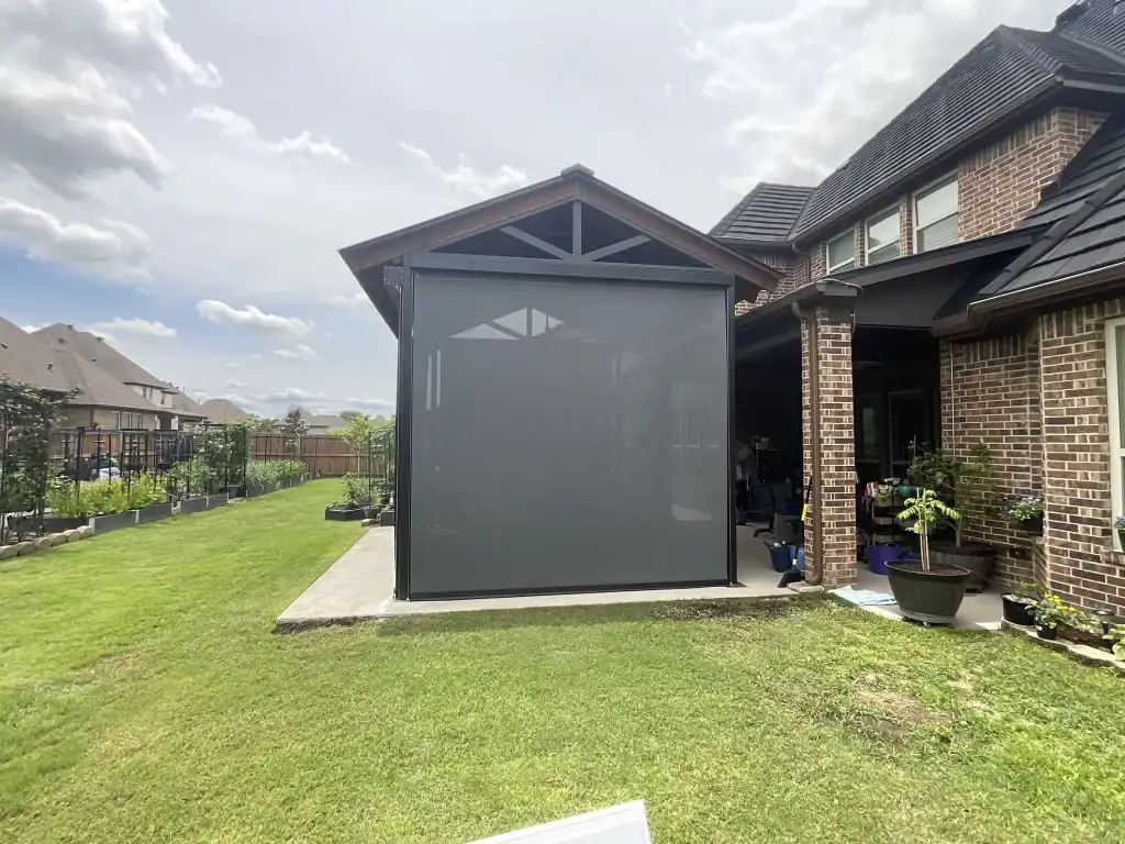 A patio of a brick house with a covered extension featuring a large gray retractable screen, surrounded by a green lawn and garden beds under a cloudy sky.