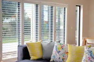 Elegant white plantation shutters on large window with colorful throw pillows on sofa.