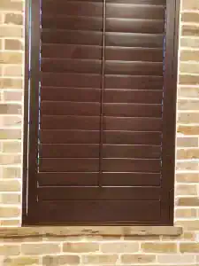 A closed wooden shutter with horizontal slats is set in a brick wall. The dark brown shutter contrasts with the light-colored bricks surrounding the window frame.