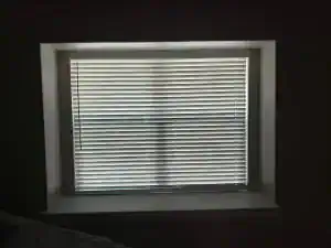 A window with closed white horizontal blinds is set in a recessed frame. Sunlight filters through the blinds, casting faint shadows in a cross pattern on the surface. The room appears dimly lit.