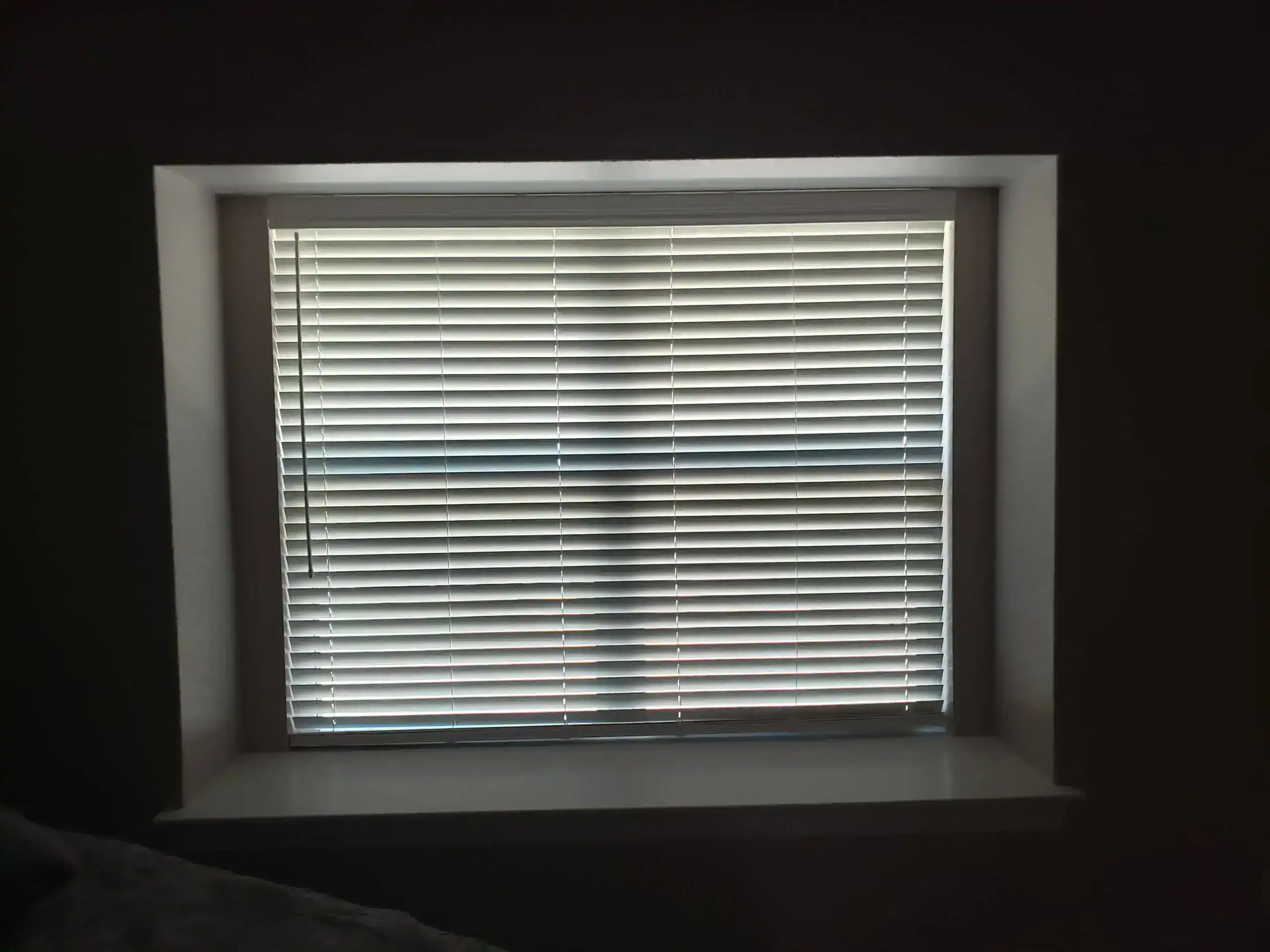A window with closed white horizontal blinds is set in a recessed frame. Sunlight filters through the blinds, casting faint shadows in a cross pattern on the surface. The room appears dimly lit.