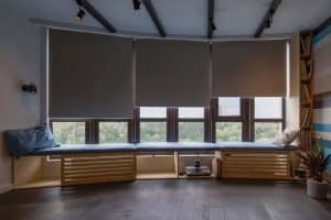 A cozy modern window seat with blue cushions, built-in wooden benches, and large windows covered by roller shades; books and decor are neatly arranged on nearby shelves.