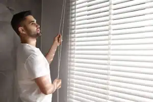 A man in a white t-shirt stands indoors, pulling the cord of white window blinds, letting sunlight into the room.