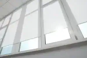 Large windows with white roller blinds partially drawn, letting in soft sunlight. The scene is bright, minimal, and modern, with clean lines and a white interior.