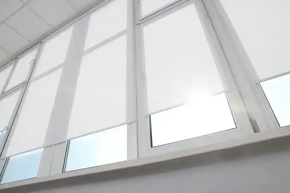 Large windows with white roller blinds partially drawn, letting in soft sunlight. The scene is bright, minimal, and modern, with clean lines and a white interior.