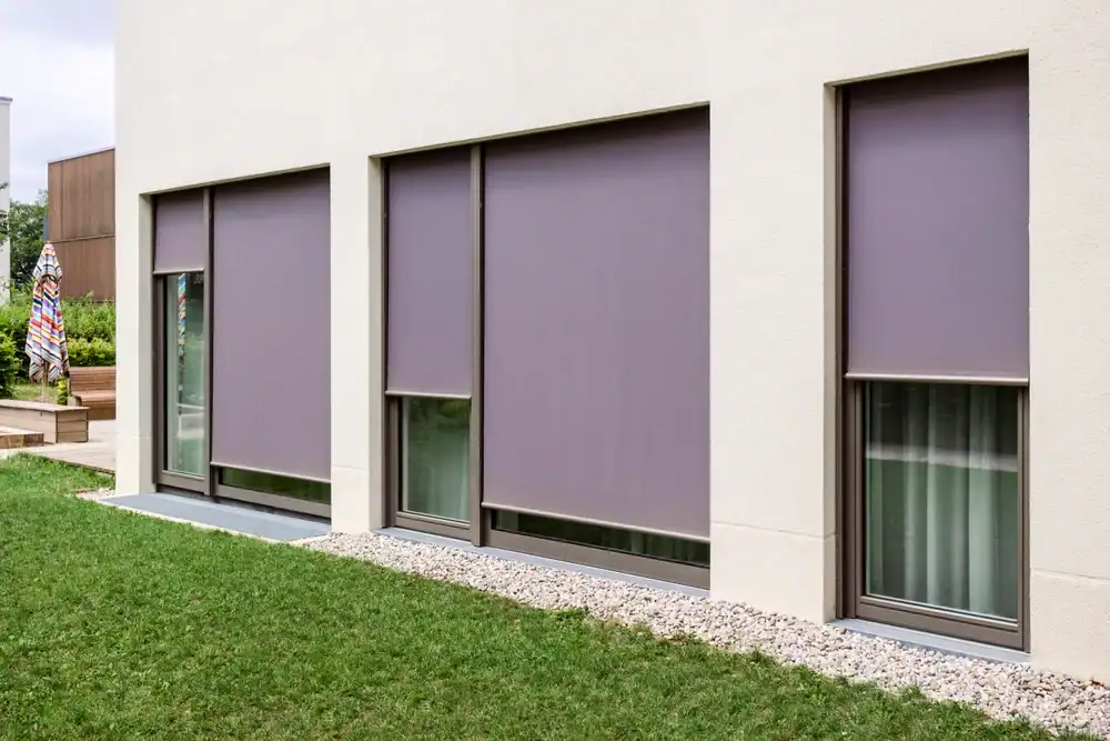 Three large windows with closed gray roller blinds on a modern white building, with a strip of white stones at the base and green grass in the foreground.